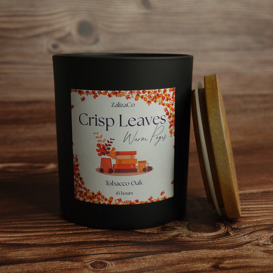 A black candle jar labeled 'ZalizaCo Crisp Leaves Warm Pages Tobacco Oak 45 hours' sits on a rustic wooden surface with its wooden lid leaning against it.