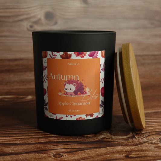 A matte black candle jar with a wooden lid leaning against it on a rustic wood surface. The front label is orange with autumn-themed illustrations and text reading 'Autumn Bliss Apple Cinnamon' and 'ZalizaCo'.