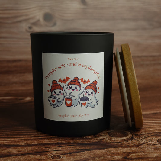 A black candle jar with a white label featuring three cartoon ghosts in orange beanies and red cups, next to its wooden lid on a rustic wooden table. The label reads 'ZaltzoCo pumpkin spice and everything nice' and 'Pumpkin Spice / Soy Wax'.