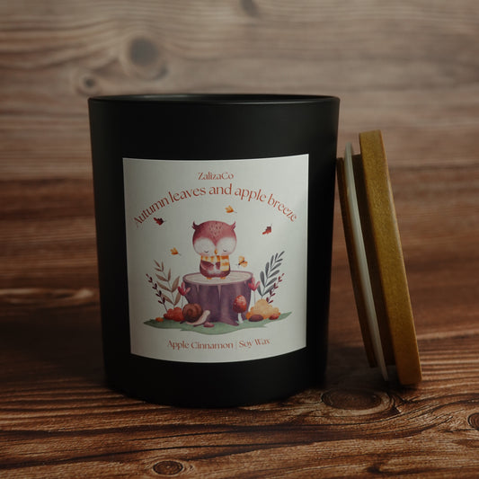 A black candle jar with a white illustrated label reading 'Autumn leaves and apple breeze' and 'ZalicaCo', shown on a wooden surface with its lid leaning beside it.