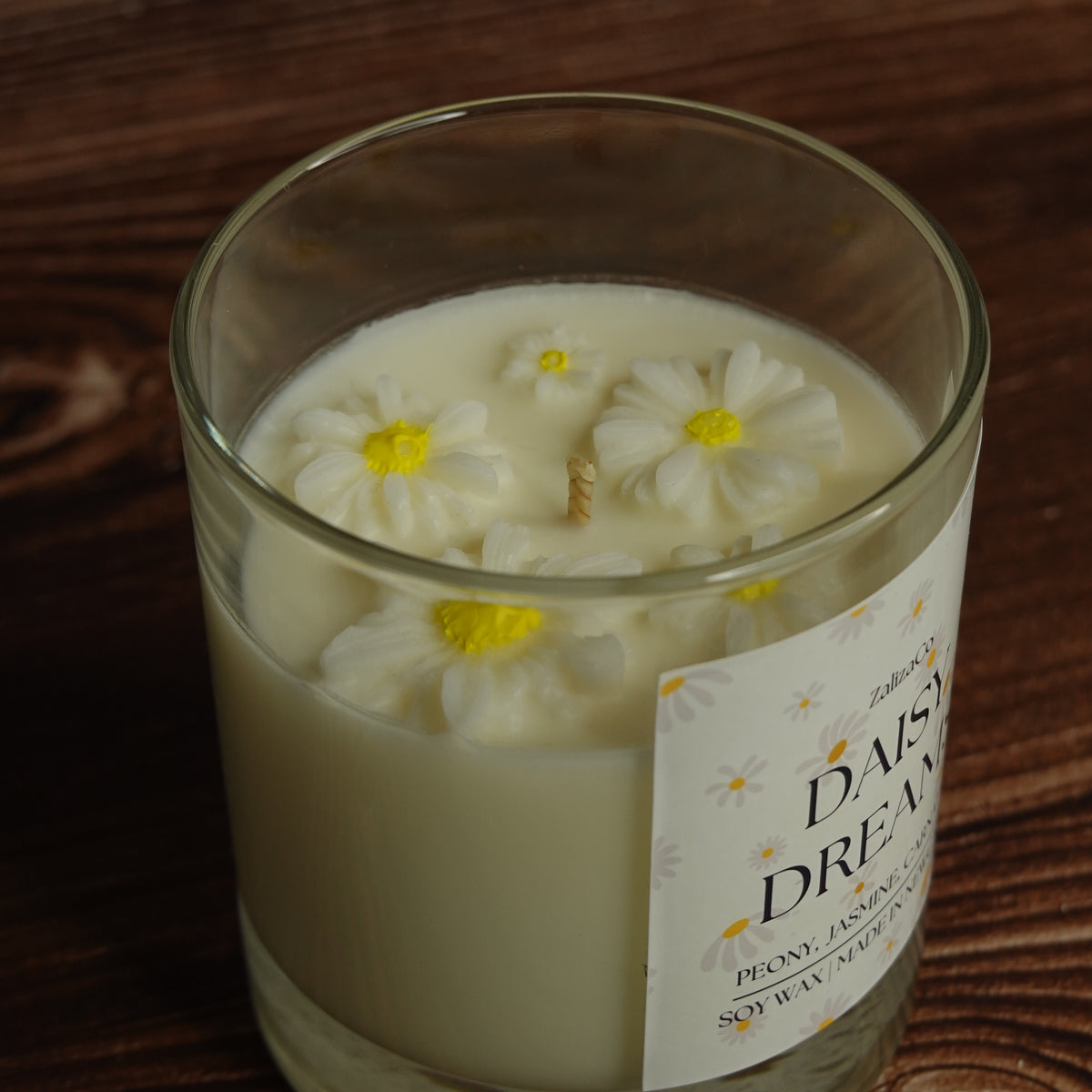 A clear glass jar candle filled with creamy off-white wax and decorated with three white daisy flowers with yellow centers. The candle has a white label reading 'DAISY DREAMS' and is photographed on a dark wooden surface.