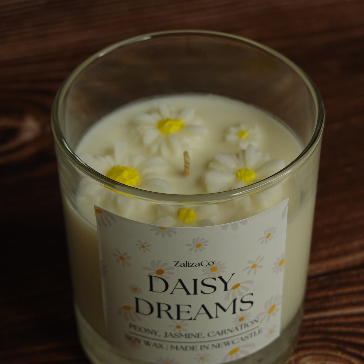A glass jar candle labeled 'Daisy Dreams' by ZalizaCo, filled with creamy off-white wax and decorated with three 3D daisy flowers on top, resting on a dark wooden surface.