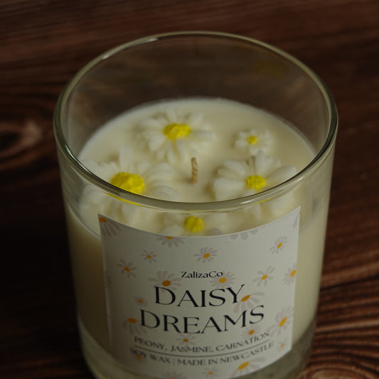 A glass jar candle labeled 'Daisy Dreams' by ZalizaCo, filled with creamy off-white wax and decorated with three 3D daisy flowers on top, resting on a dark wooden surface.