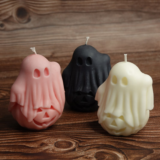 Three ghost-shaped candles in pink, black, and cream, arranged on a rustic wooden surface.