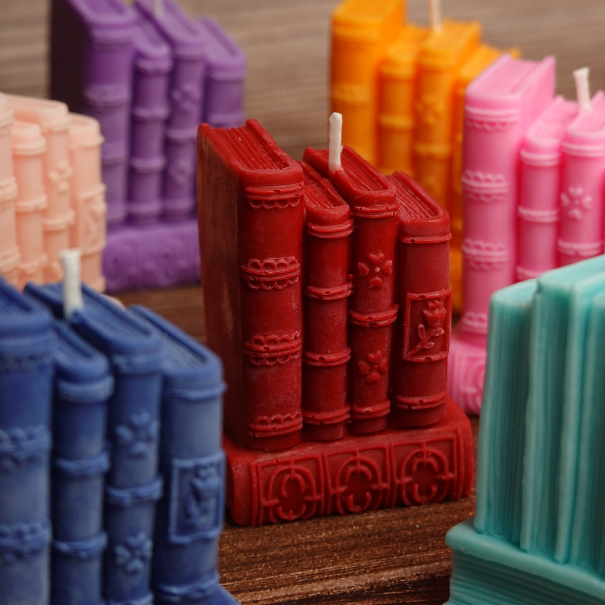 A close-up of a novelty candle shaped like a row of old book spines, with wicks sticking out. The main candle is red, but other coloured book candles in blue, purple, orange, pink, and teal are visible in the background and foreground on a wooden surface.