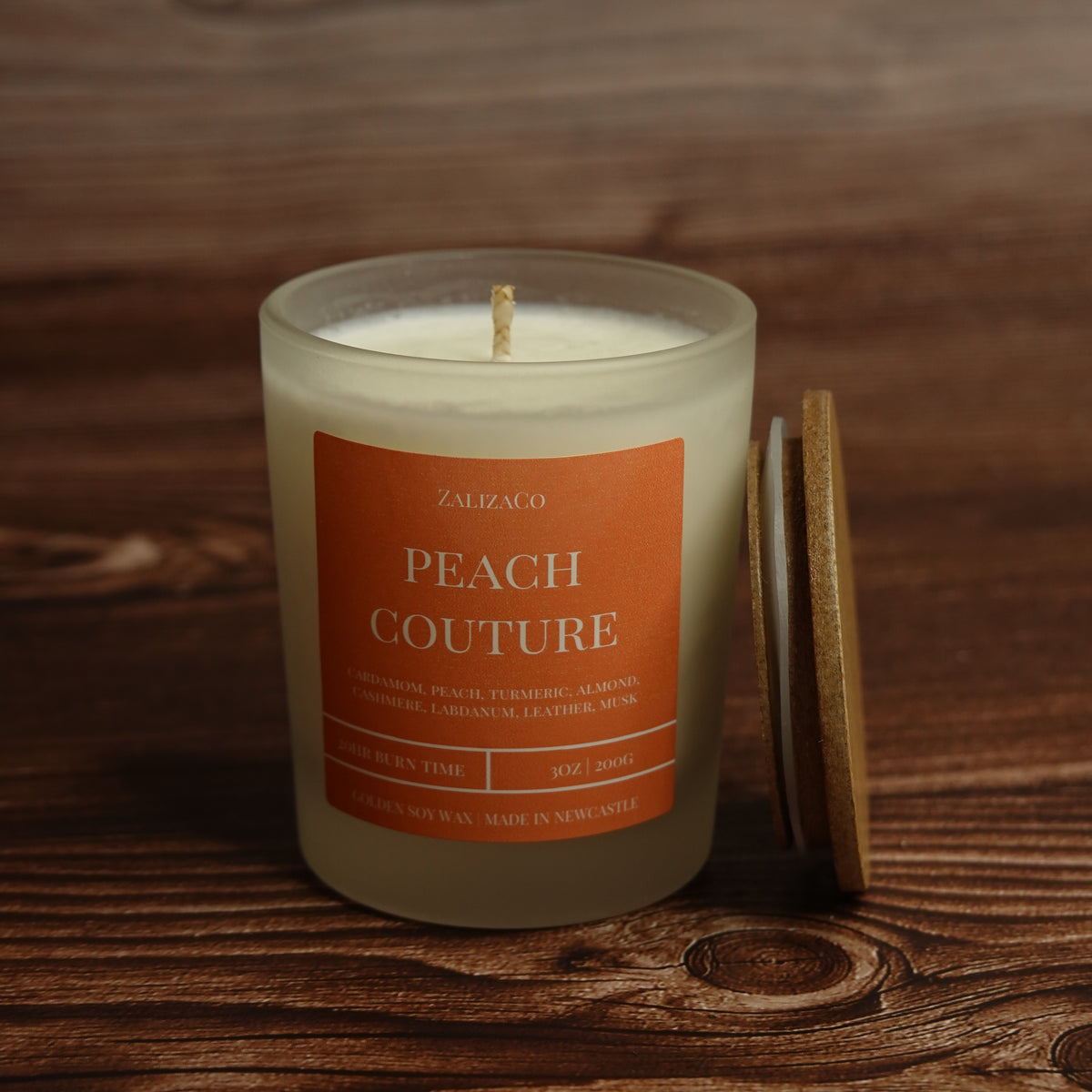 A frosted glass candle jar labeled 'ZALIZACO PEACH COUTURE' sits on a dark wooden surface with its wooden lid leaning against it. The candle wax is creamy off-white, and the label is a warm peach-orange color.