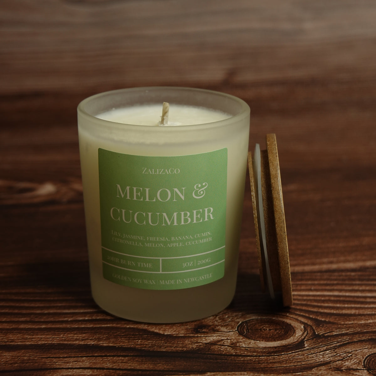 A glass jar candle labeled 'ZALIZACO MELON & CUCUMBER' sits on a dark wooden surface, with a wooden lid leaning against the right side of the jar.