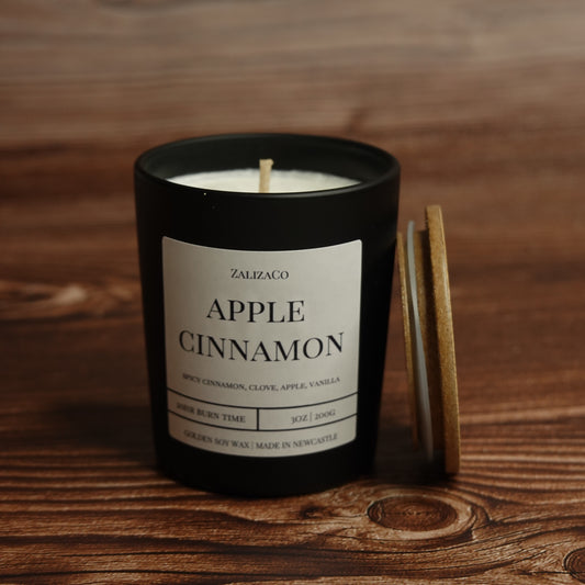 A black glass jar candle with a white label reading 'ZALIZACO APPLE CINNAMON' sits on a wooden surface, with a light wood lid leaning against it.