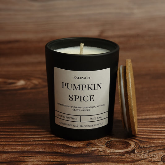 A black candle jar labeled 'ZALIZAGO PUMPKIN SPICE' with cream-colored wax and a wooden lid resting beside it on a rustic wooden tabletop.