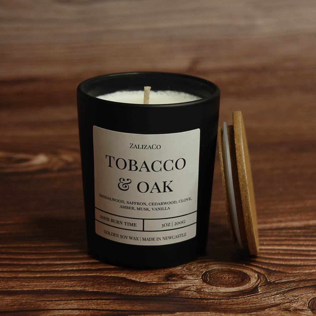 A black glass candle jar with a white label reading 'TOBACCO & OAK' by ZalizaCo, placed on a wooden surface with a wooden lid leaning against it.