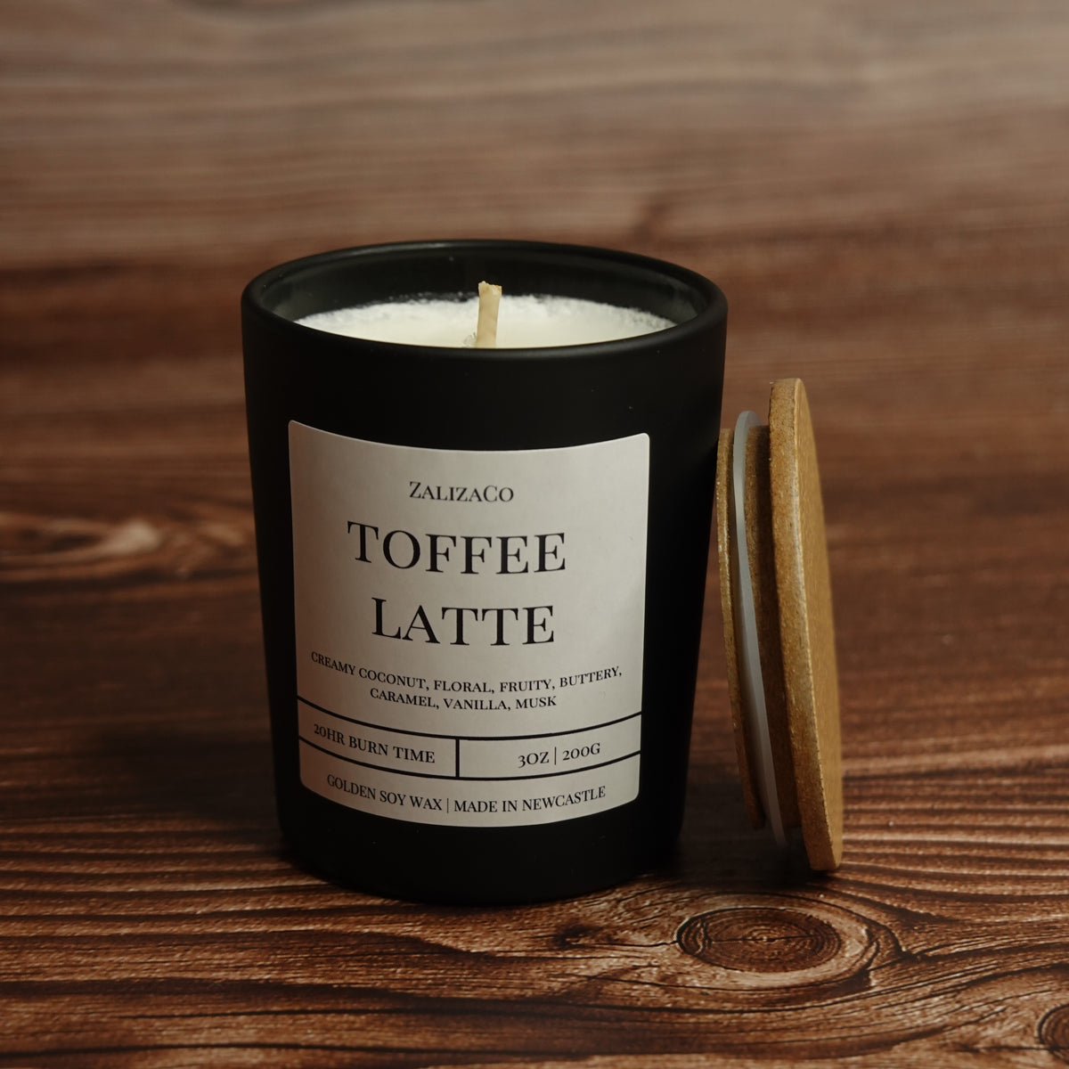 A black glass candle jar labeled 'ZALIZACO TOFFEE LATTE' with a white wax wick visible, set on a wooden surface beside a light wood lid.