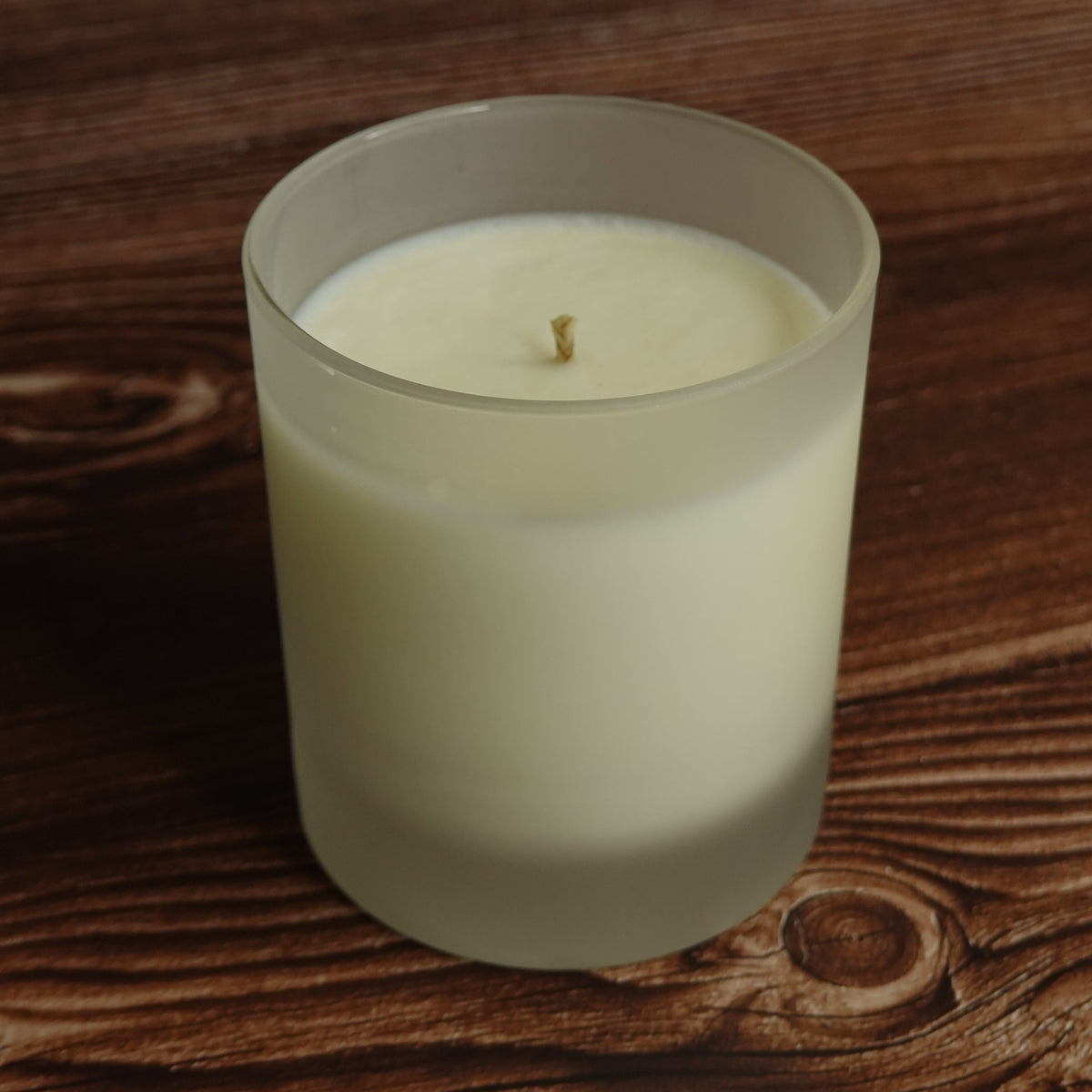 A frosted glass jar candle with creamy off-white wax and a single wick, set on a dark brown wooden surface.