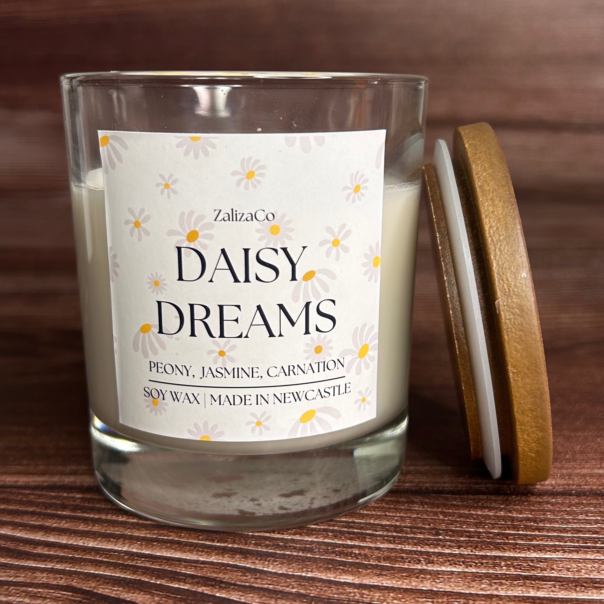 A clear glass jar candle with white wax and a white label reading 'Daisy Dreams' by ZalizaCo, shown on a wooden surface with a round wooden lid leaning against the jar.